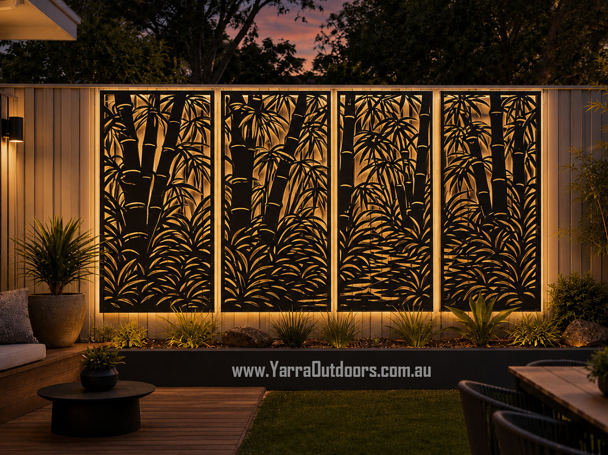 A decorative outdoor wall panel set with a black bamboo leaf pattern, illuminated from within to show a warm golden glow against a wooden fence in a backyard setting.