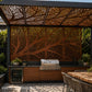 Outdoor kitchen setup under a decorative laser-cut metal pergola with a tree branch pattern, featuring a stainless steel gas grill, a wine cooler, and a wooden dining table with chairs.