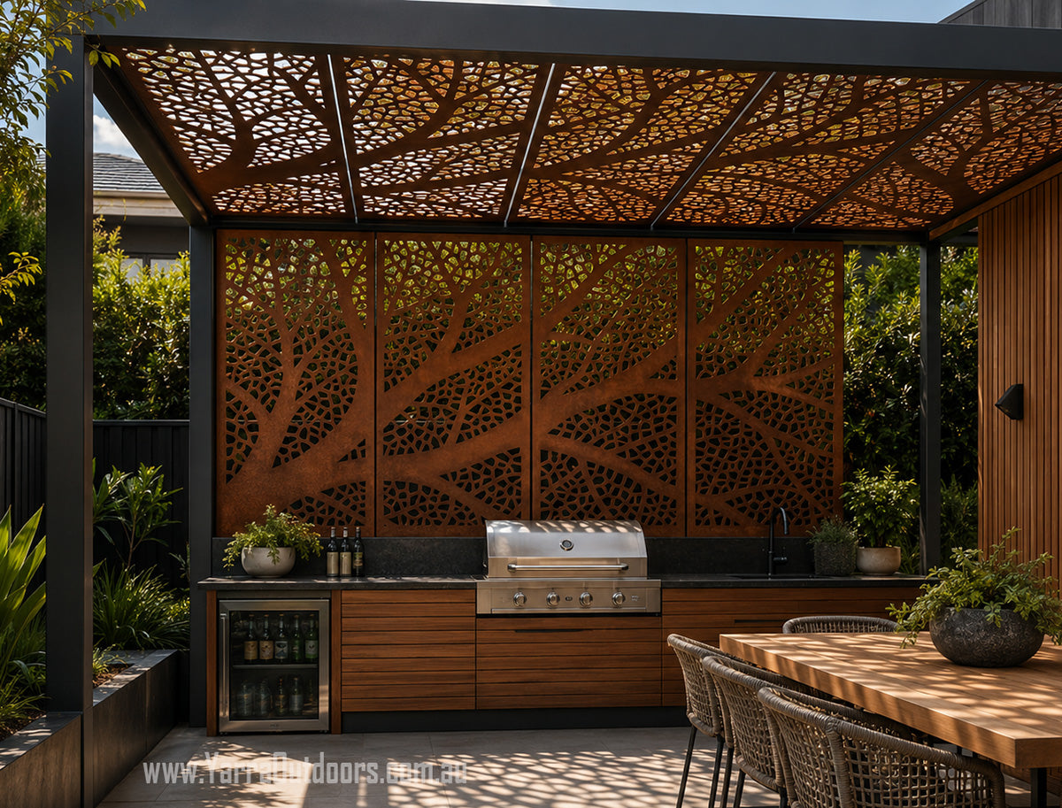 Outdoor kitchen setup under a decorative laser-cut metal pergola with a tree branch pattern, featuring a stainless steel gas grill, a wine cooler, and a wooden dining table with chairs.